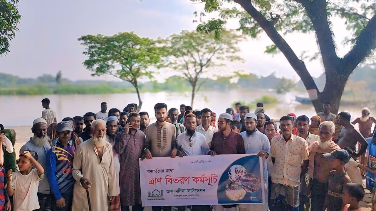 জলাভূমির পাশে একদল মানুষ বন্যার্তদের ত্রাণ বিতরণের ব্যানার ধরে দাঁড়িয়ে আছেন।
